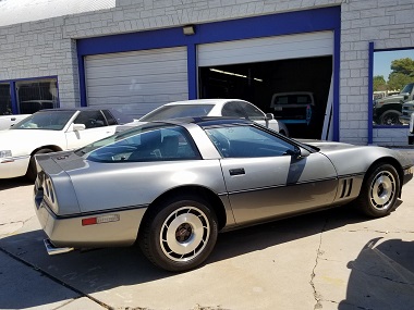 Silver Chevrolet Corvette C4 parked in front of the Denvers Car Care garage with other cars. In the background, the service bay door is open and there is a pickup truck inside the garage.
